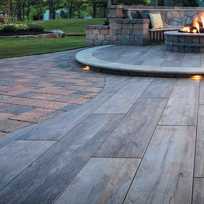 Wooden patio with curved edge, leading to fire pit and stone bench. Grass and trees in background.