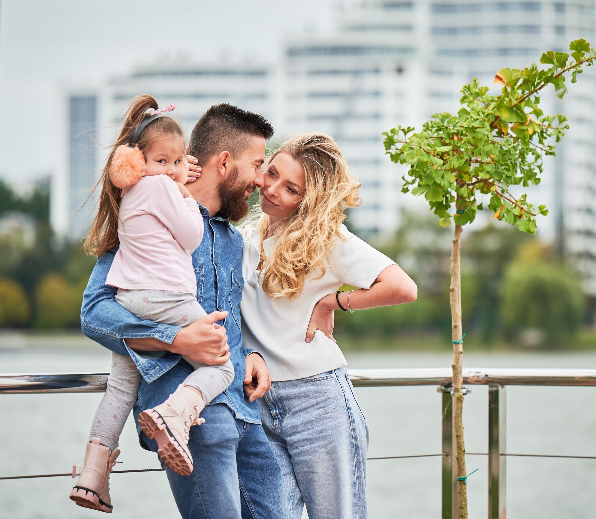 Family of three embracing near a waterfront; child in arms, adults smiling, cityscape background.