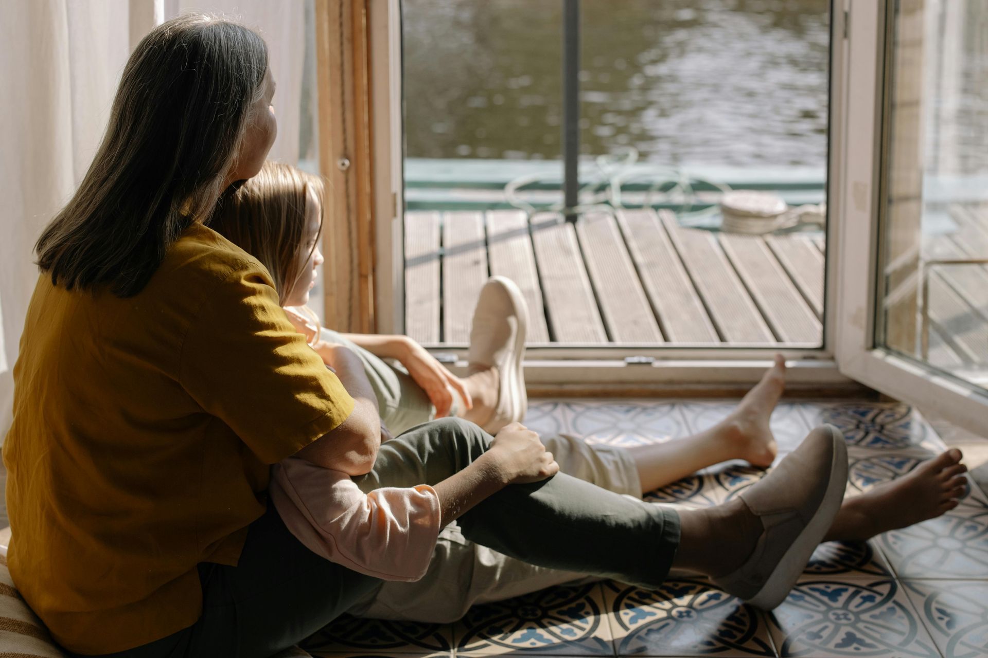 Two people sitting together on a tiled floor by an open door, looking out at a body of water and a wooden deck.