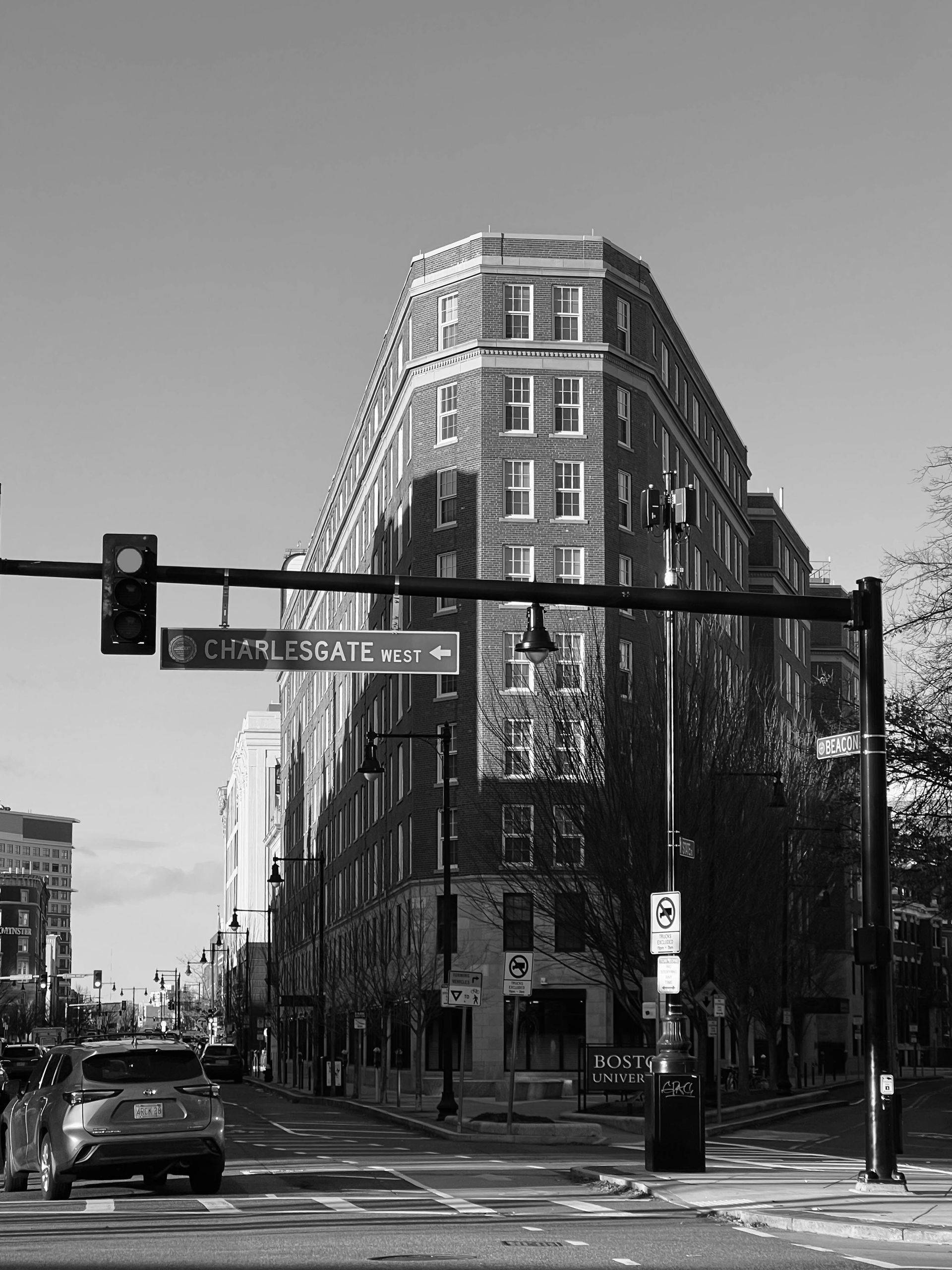 A black-and-white view of a wedge-shaped brick building at a city intersection, featuring a street sign and a car.