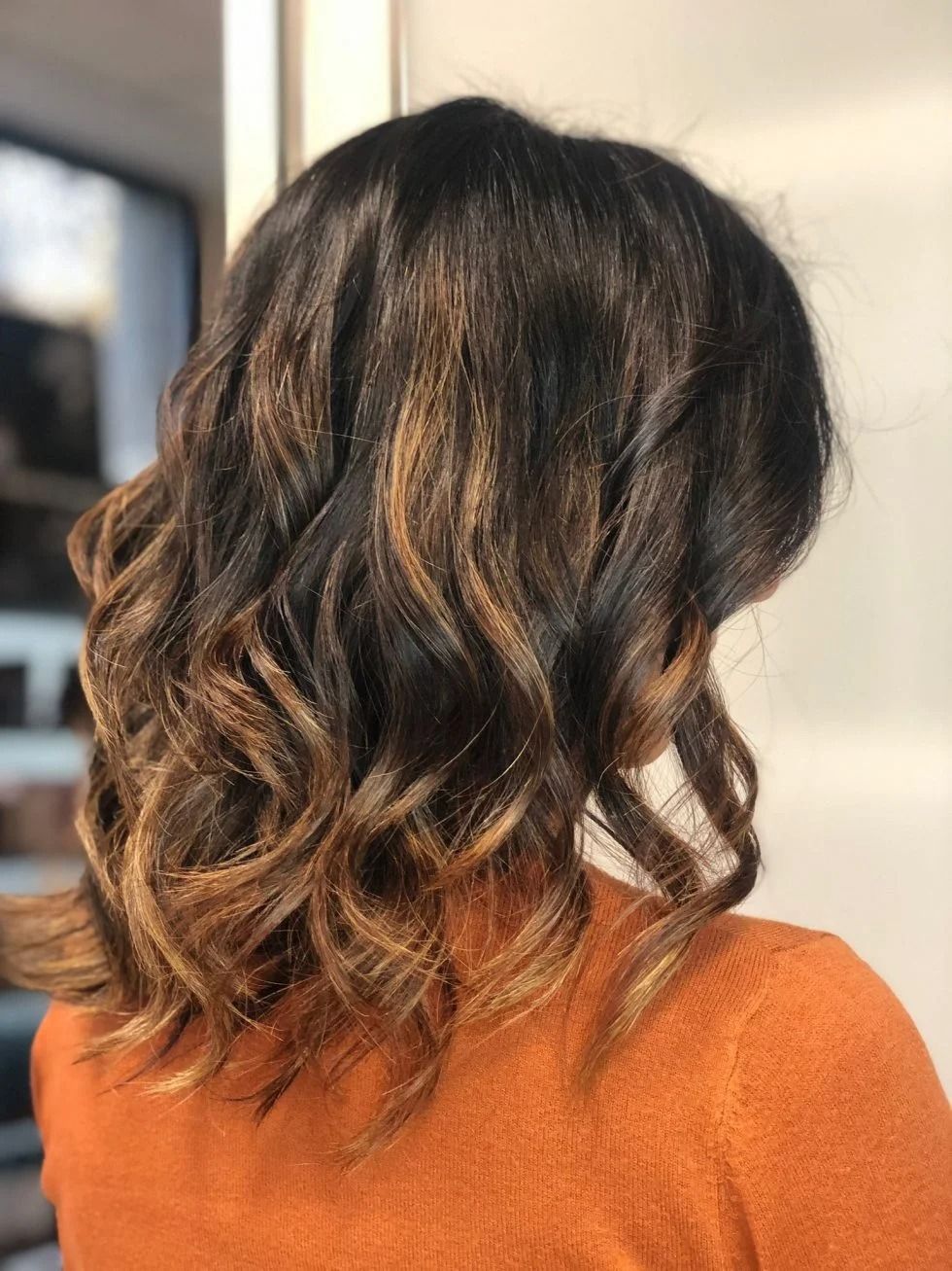 Donna con capelli castani lunghi fino alle spalle, acconciati con onde morbide e riflessi dorati. Indossa un maglione arancione.