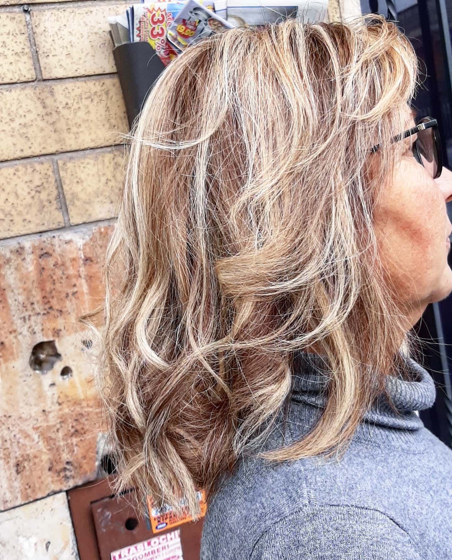 Donna con capelli biondi e castani ondulati e scalati, occhiali da sole e dolcevita grigio. Sfondo muro di mattoni.