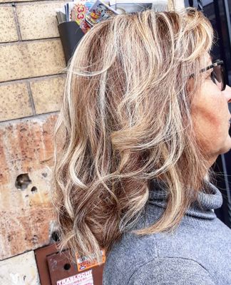 Donna con capelli biondi e castani ondulati e scalati, occhiali da sole e dolcevita grigio. Sfondo muro di mattoni.