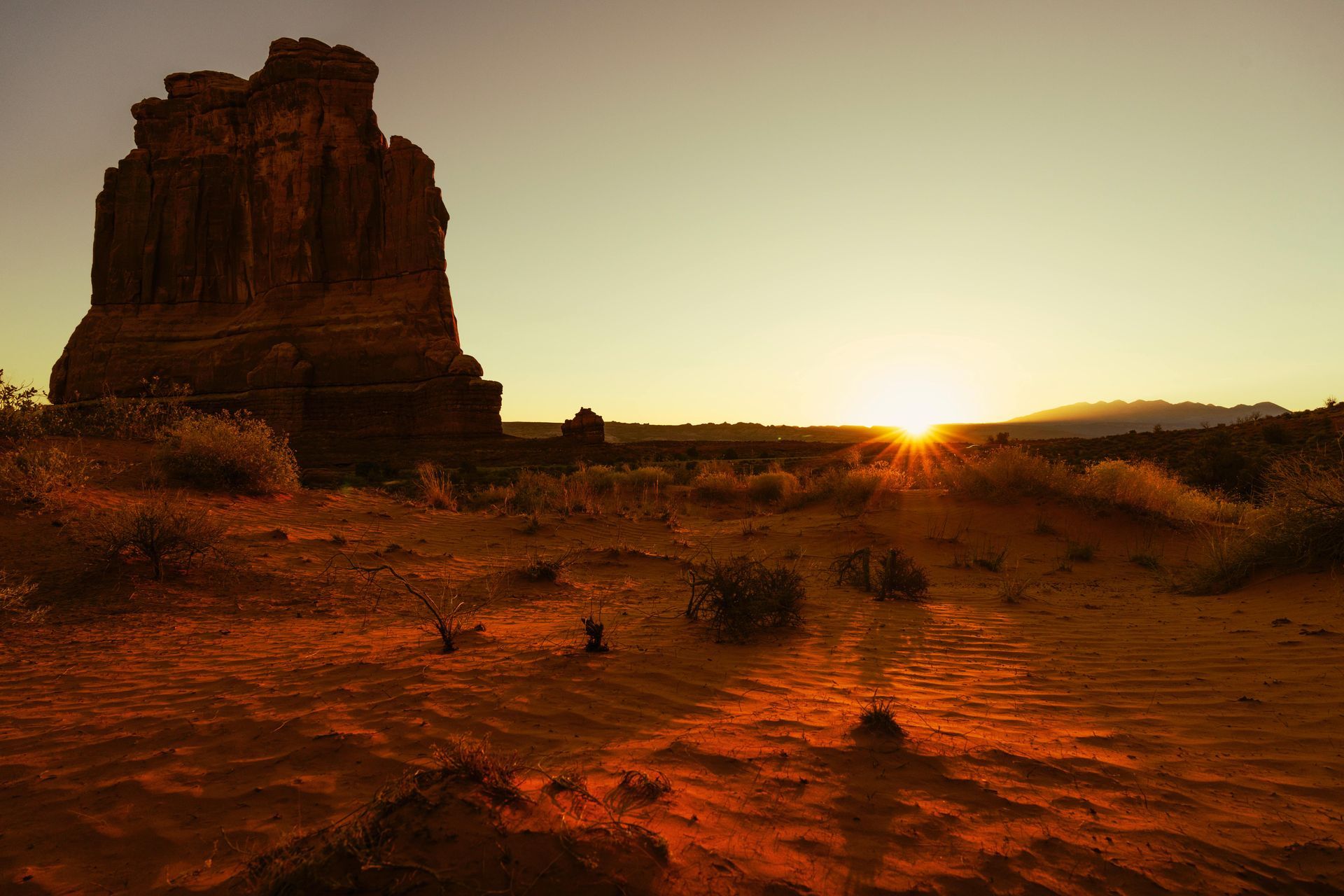 The sun is setting over a desert landscape with a large rock formation in the background.