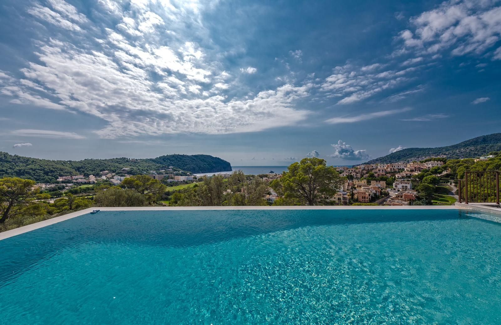 Infinity pool with turquoise water overlooking a town, trees, and ocean under a cloudy sky.