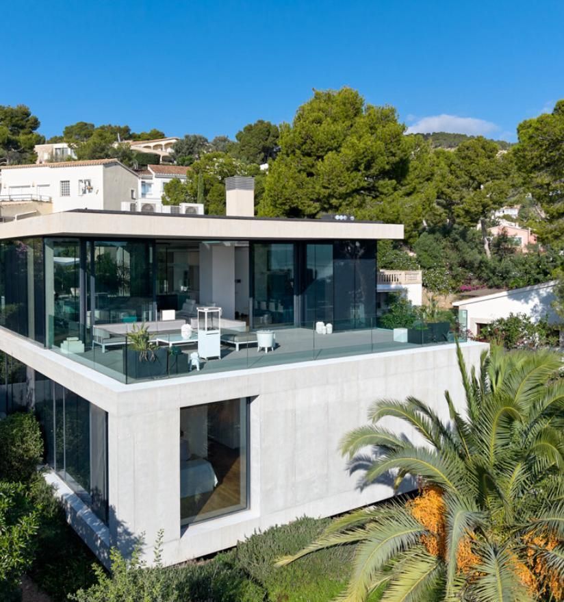 Modern, two-story house with glass walls and a rooftop terrace, surrounded by palm trees and greenery on a sunny day.