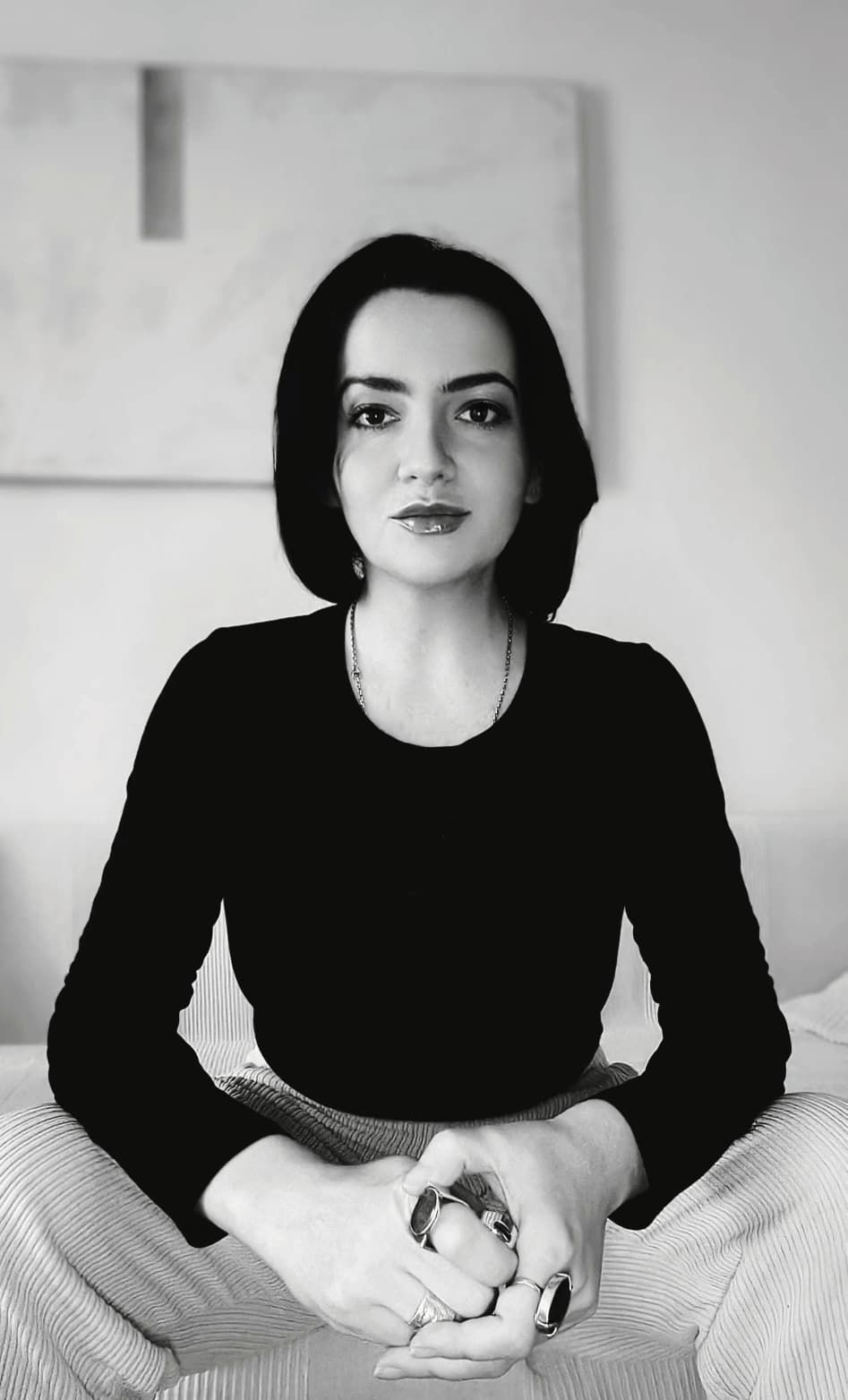 Woman seated, wearing a black shirt and light-colored pants, hands clasped, looking at the camera.