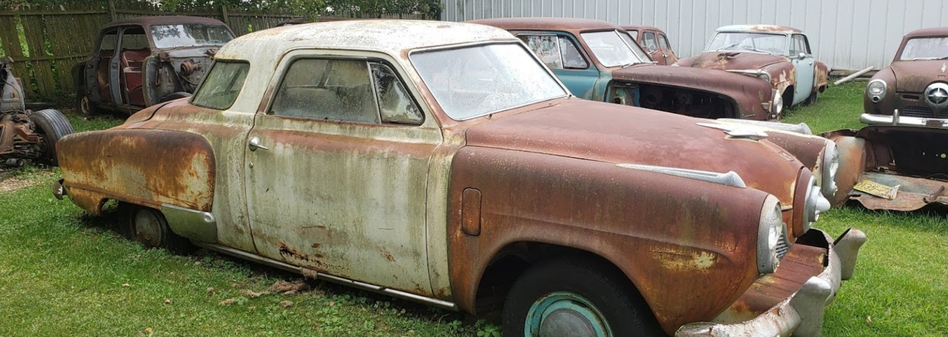 A row of old rusty cars are parked in the grass.
