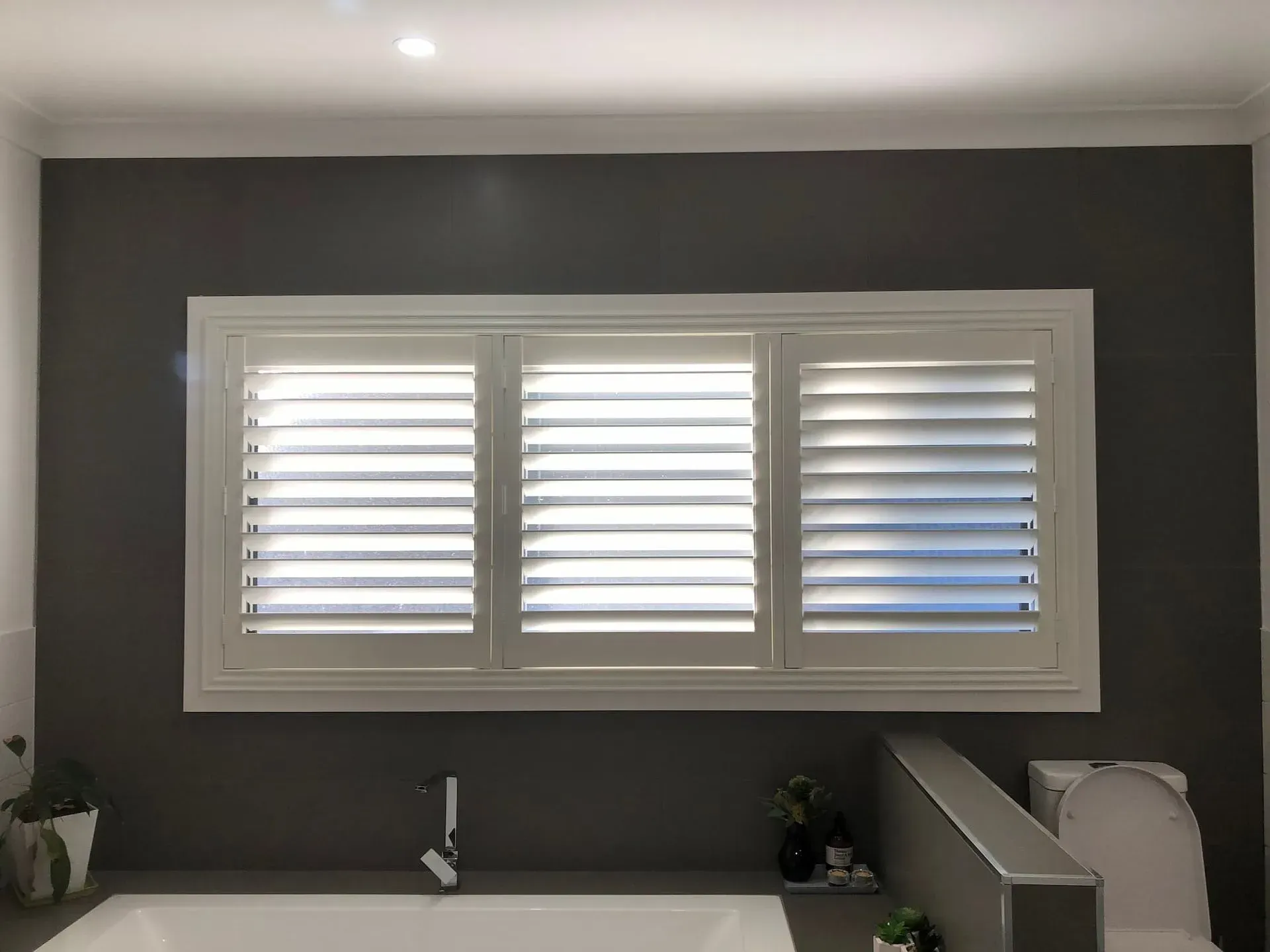 White window shutters in a gray-walled bathroom above a bathtub, with a sink and toilet visible.