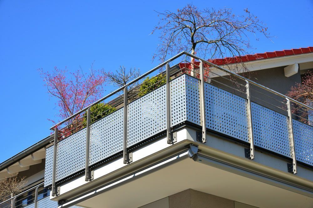 Modern balcony with gray perforated metal railing, plants, and a tree against a blue sky.