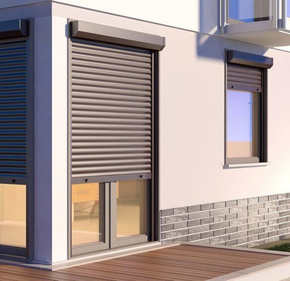 Exterior of a house with two windows and a door. Each has a closed, brown roller shutter.