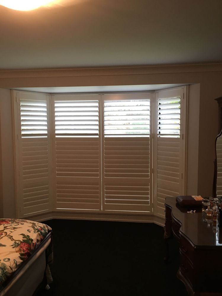 White shutters on a bay window in a bedroom, partially open, letting in light.