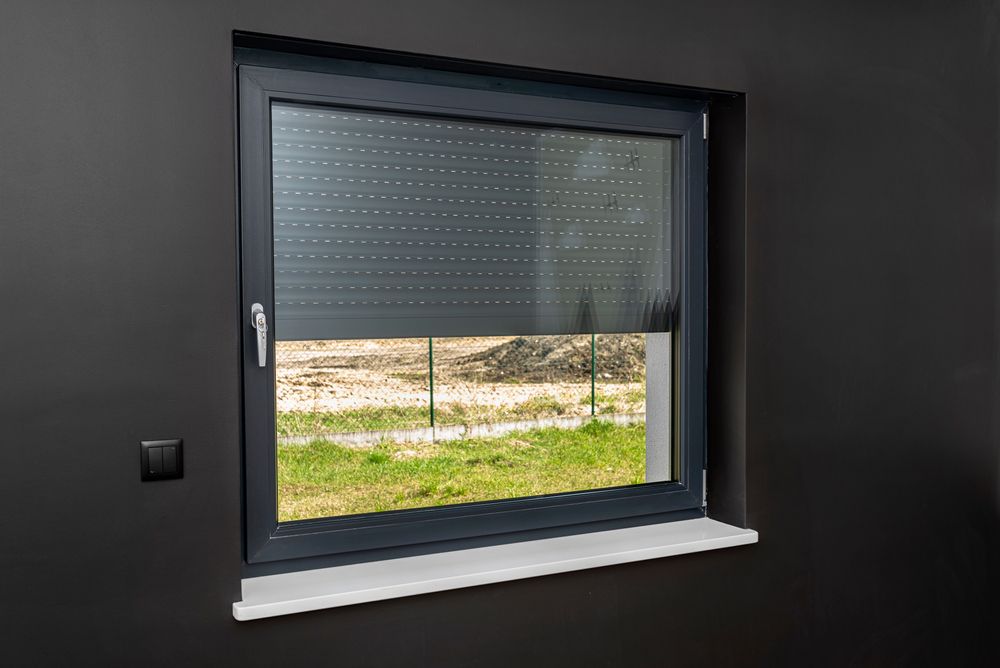 Window with dark gray frame and partially lowered gray rolling shutter, overlooking a grassy yard.