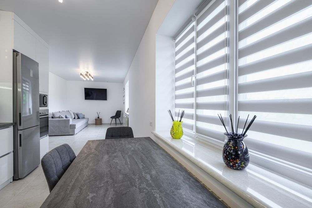 Interior view of a modern apartment with a dining table, window with blinds, and living room in the background.