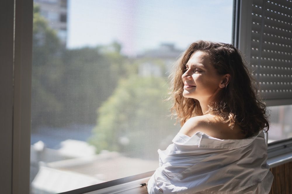 Woman smiles, looking out a window. She wears a white shirt, enjoying sunlight.