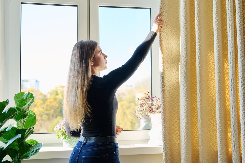 Woman pulls back a cream curtain to reveal a view from a window.