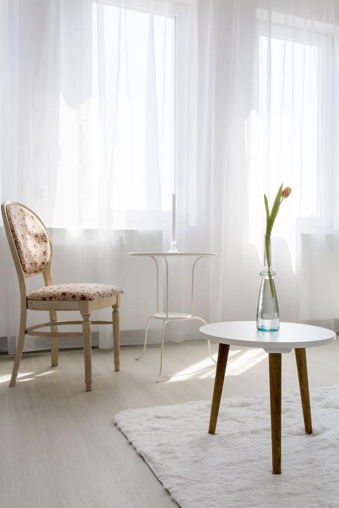 A bright, airy room with white sheer curtains, a floral-backed chair, and white tables. A vase holds a single flower.