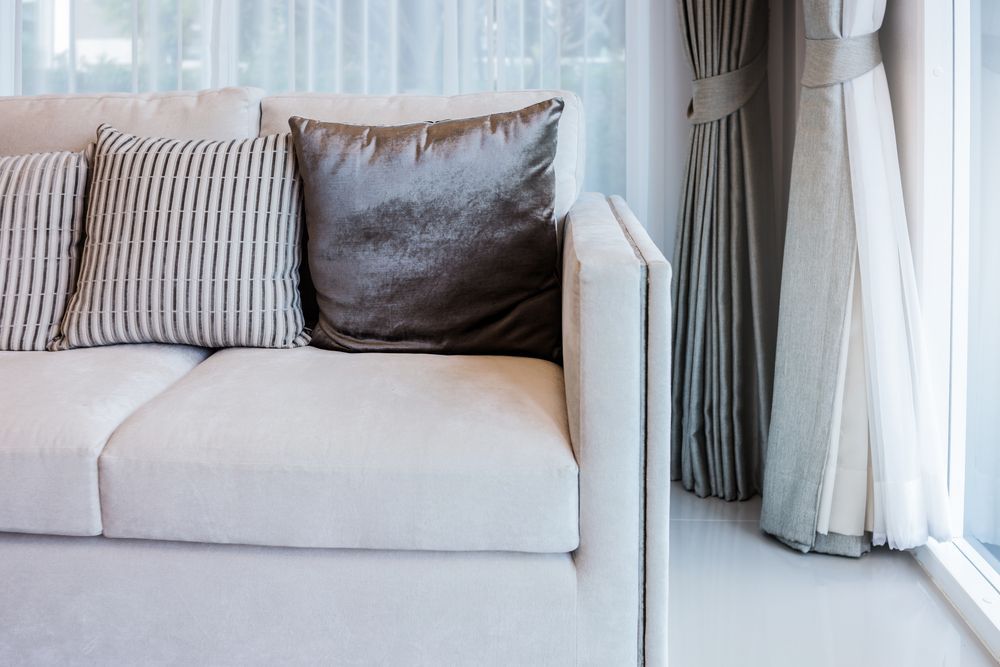 White couch with striped and velvet throw pillows, near a window with sheer curtains.