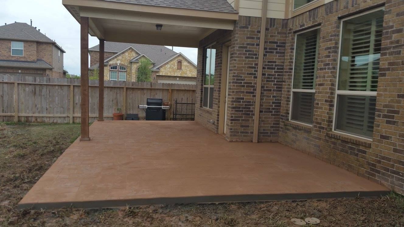 Brown concrete patio with covered overhang next to brick house; backyard setting with grill.
