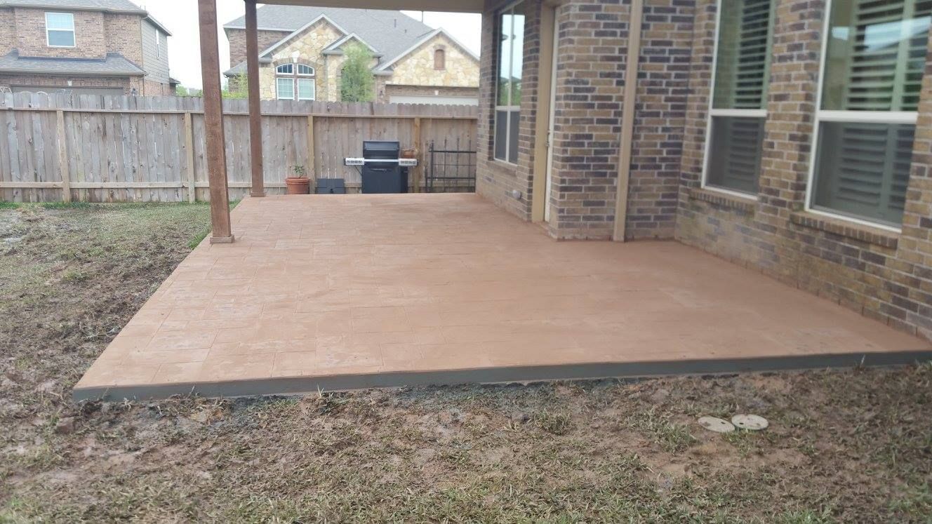 Covered concrete patio next to a brick house and grassy lawn; grill in the background.