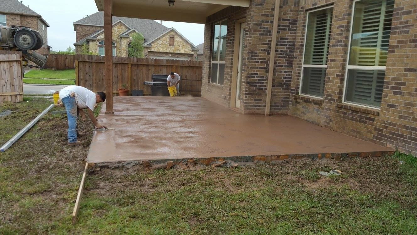 Man smoothing wet concrete patio next to a house with a covered porch and wooden fence.