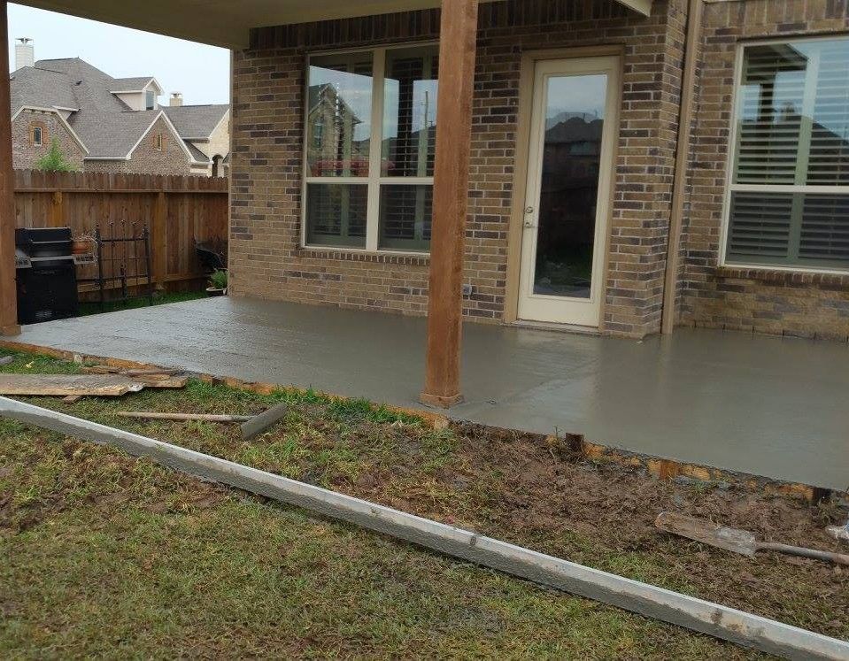 Newly poured concrete patio next to a brick house, wooden fence and green grass.