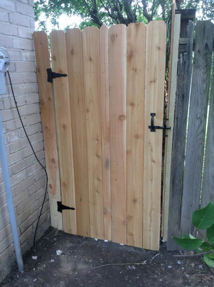Wooden gate with scalloped top, black hinges and latch, attached to brick wall and wooden fence.