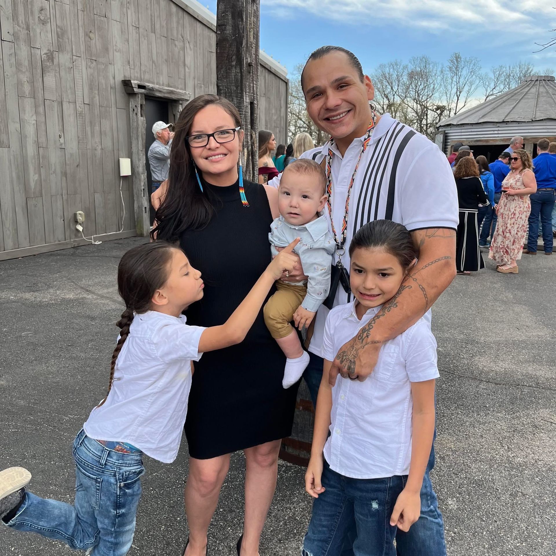 Family of five poses outdoors: woman in black dress, man holding baby, two children, smiling, light setting.