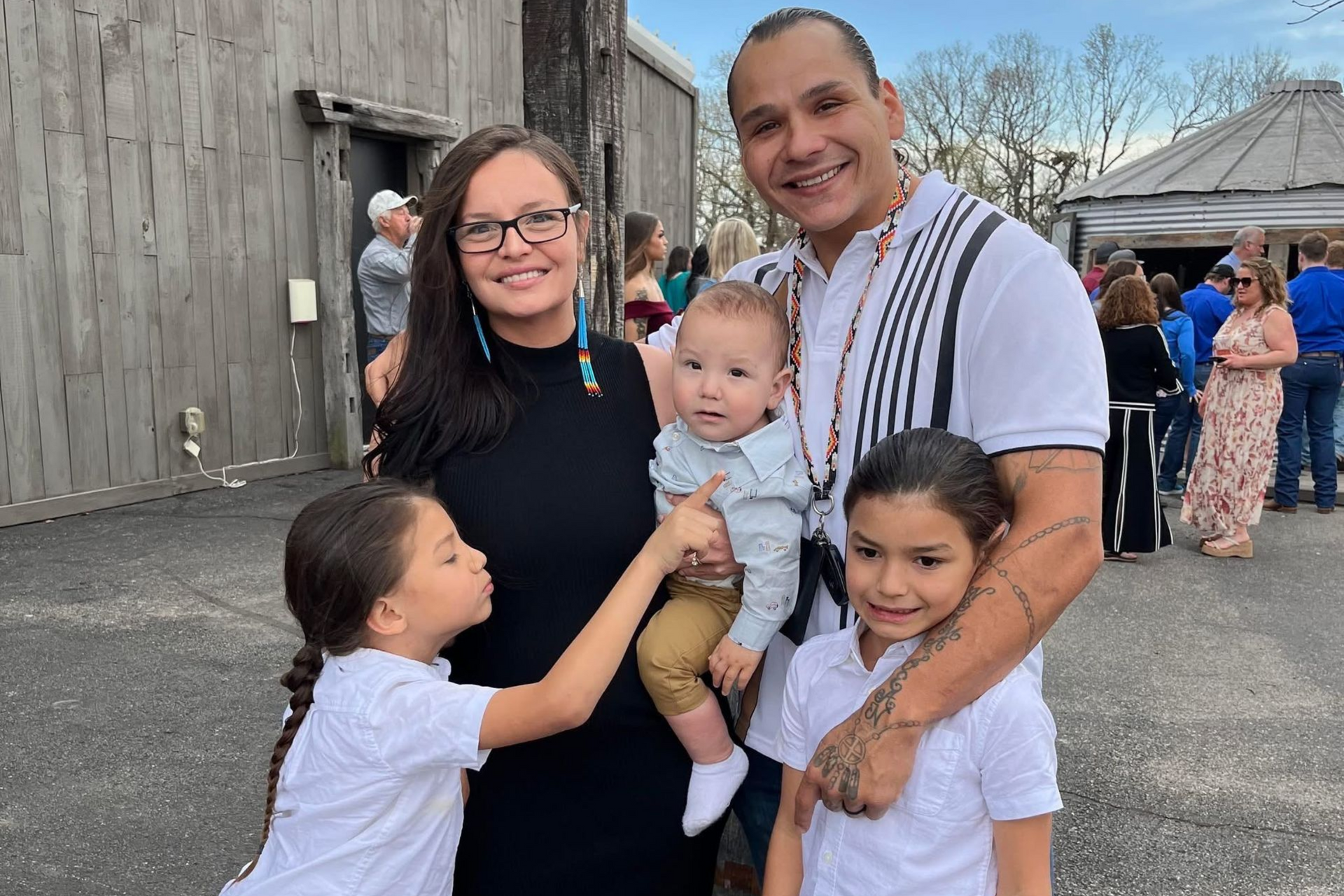 Family of five smiling outdoors; mother, father, and three children; a crowd in the background.