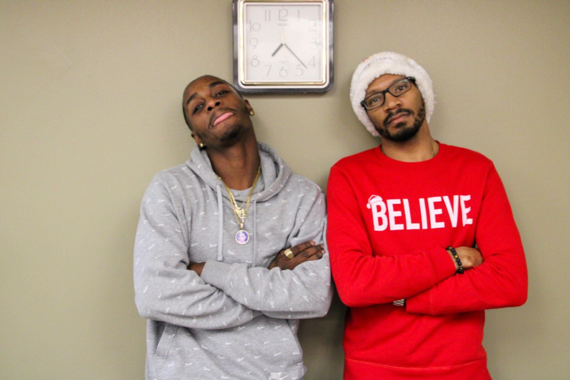 Two men are standing next to each other with their arms crossed and one is wearing a red sweater that says believe.