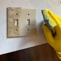 Gloved hand scrubbing a dirty light switch plate with a sponge on a wall