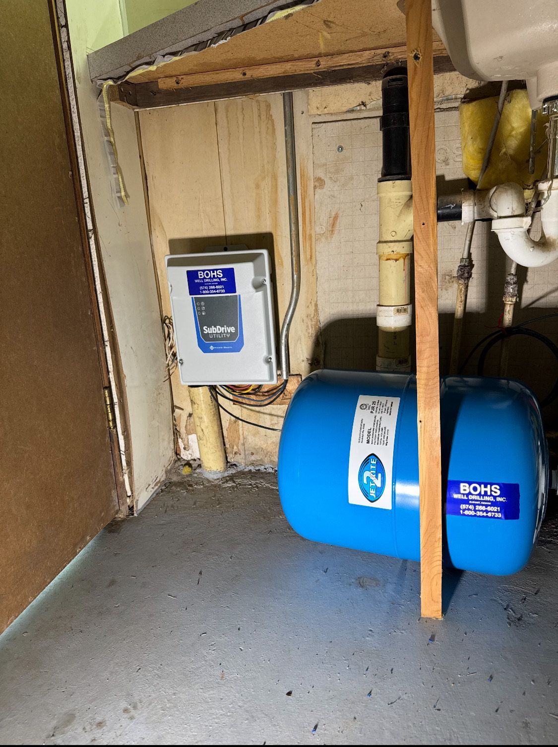 Blue water tank and control box in a basement corner. Pipes, wood framing, and concrete floor are also visible.