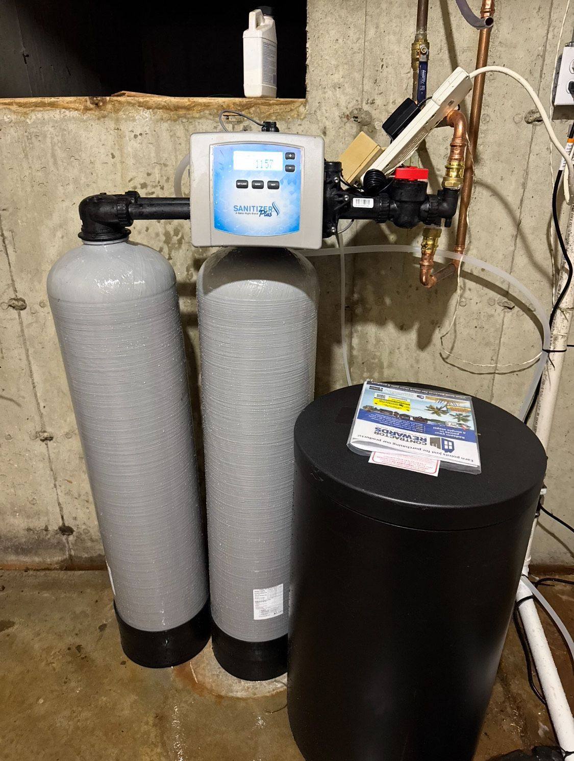 Water softening system in a basement. Includes two gray tanks, a control panel, and a black salt tank.