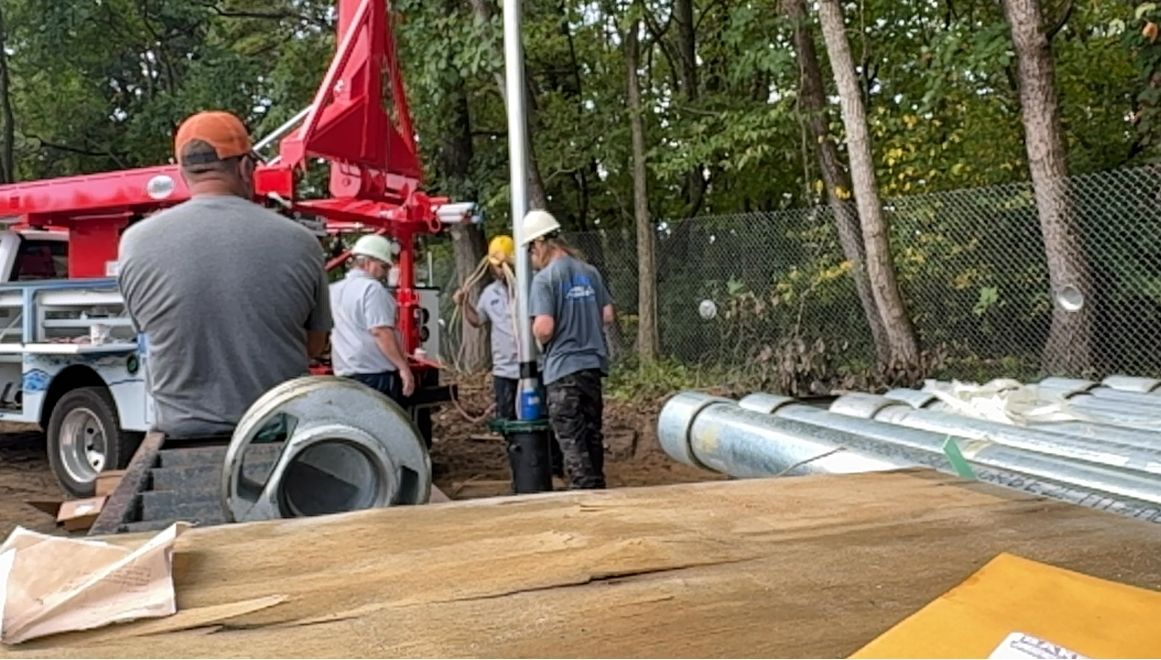 Workers installing a well. A red truck is on the left, with workers assembling pipes and equipment in a wooded area.