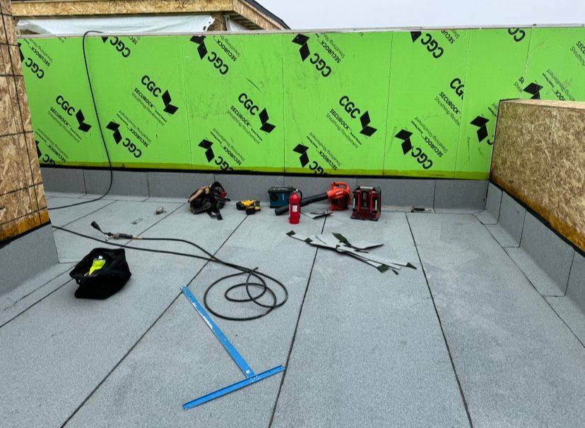 Rooftop under construction; gray surface with tools scattered. Green protective barrier and wood framing visible.