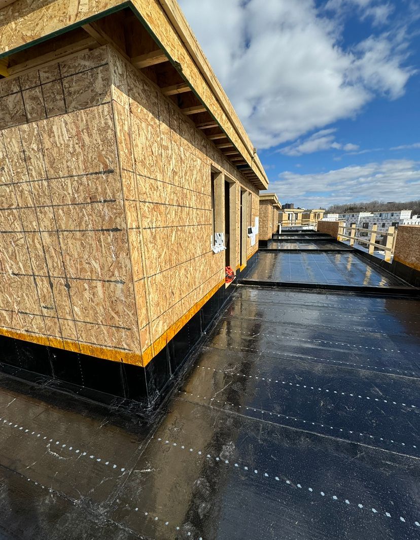 Construction site with a flat, black tar roof and unfinished wooden walls, under a cloudy blue sky.