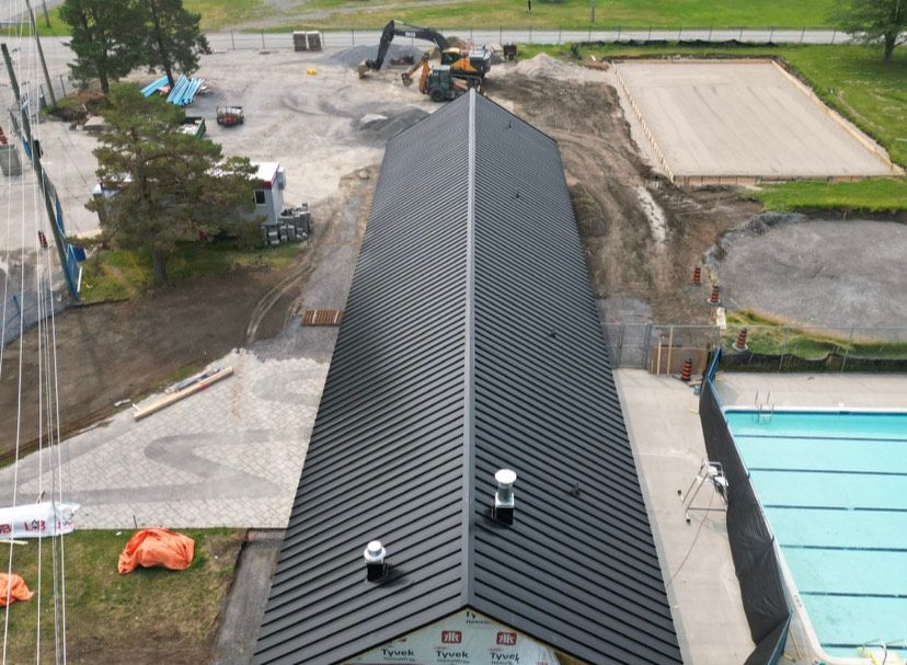 Overhead view of a long building with a black metal roof near a swimming pool. Construction equipment nearby.