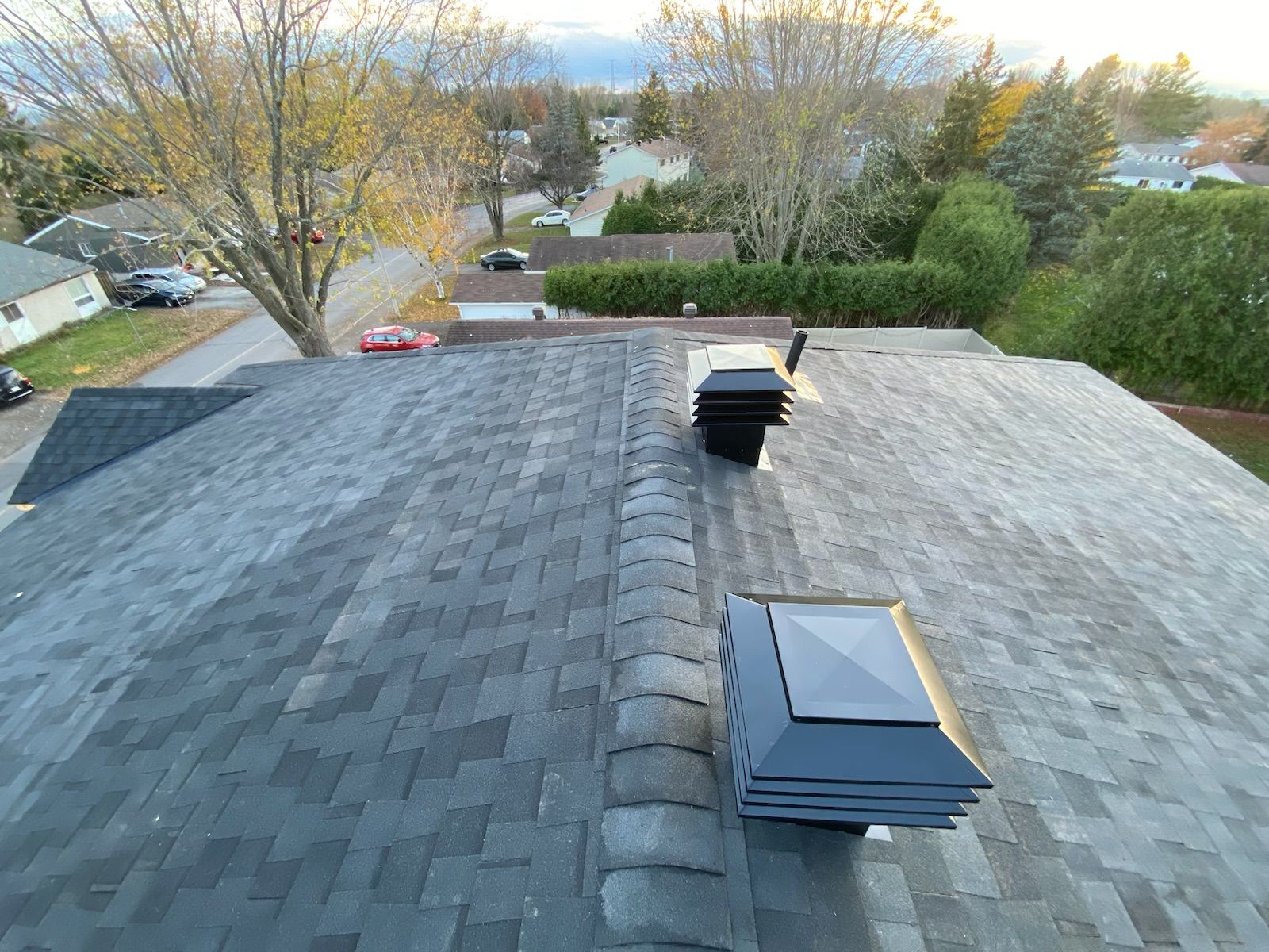 Overhead view of a gray shingled roof with skylights and vent, trees and houses in the background.