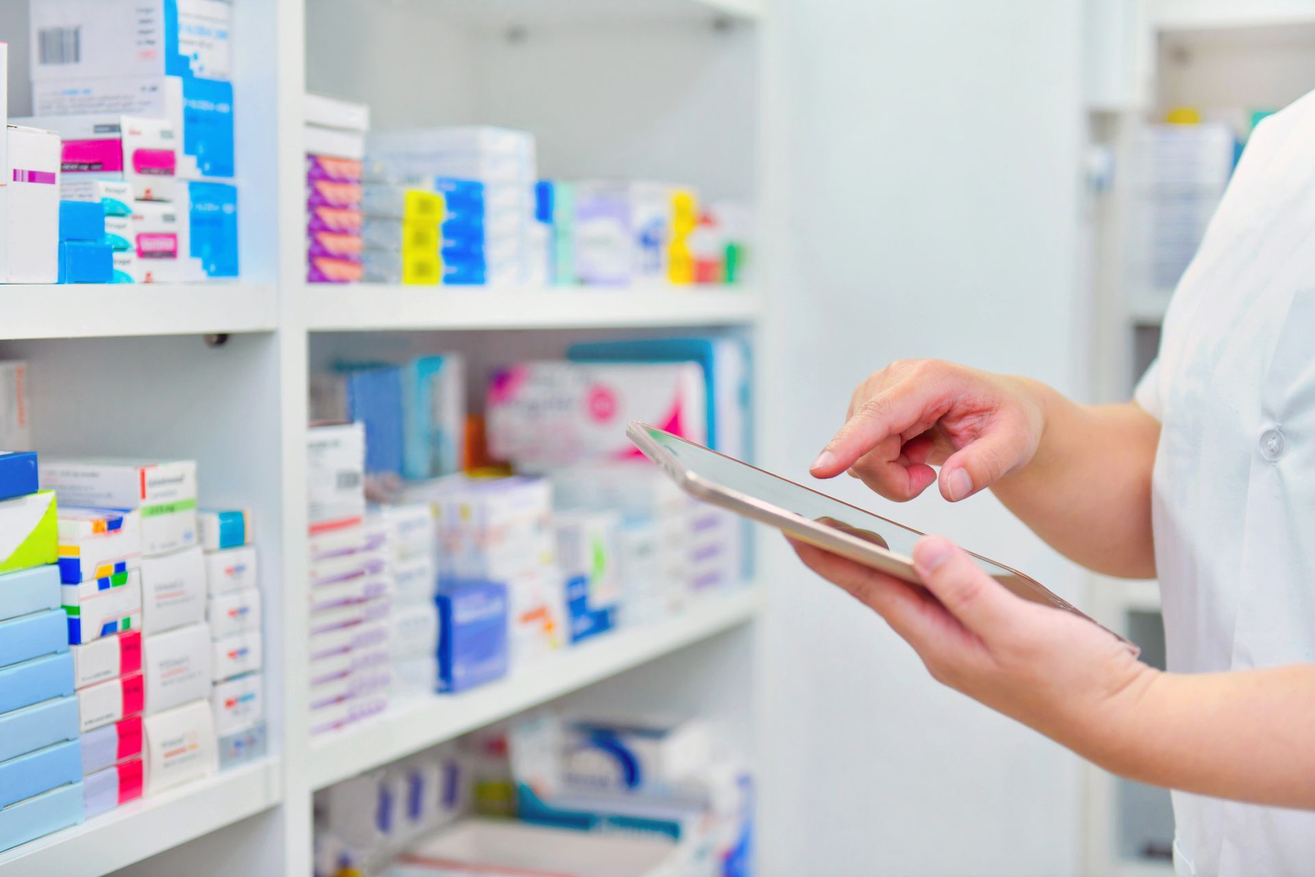 Pharmacist using tablet, checking inventory in pharmacy; shelves of medicine in background.