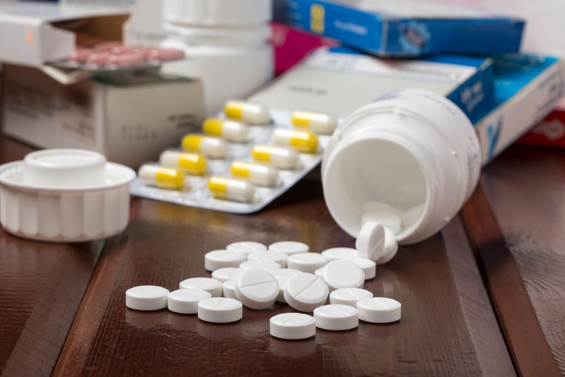 Pills spilled from a white bottle on a wooden table, surrounded by medicine bottles and packaging.