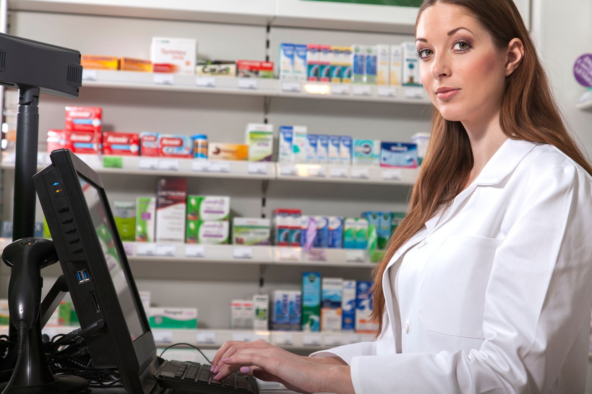 A pharmacist in a white coat works at a computer, shelves of medications in the background.