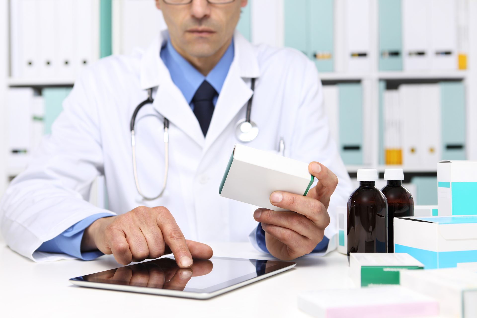 Doctor holding medication box, using a tablet. In an office setting.
