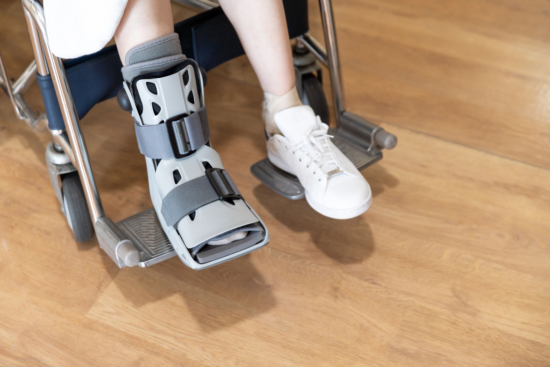 Person in wheelchair with leg brace on one foot and a sneaker on the other. Wooden floor.