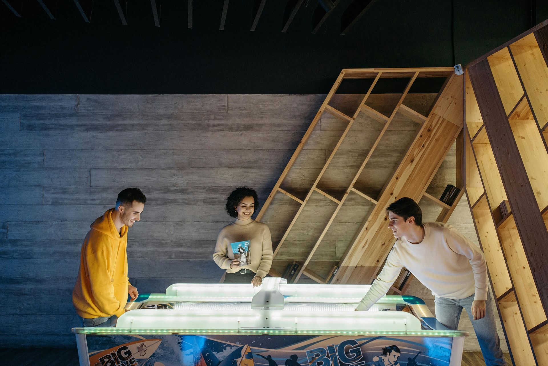 Three people playing a game at a glowing table in a room with wood and concrete walls.