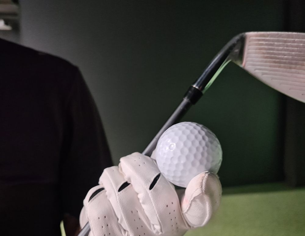 Gloved hand holding golf ball beneath club's head in front of dark backdrop.