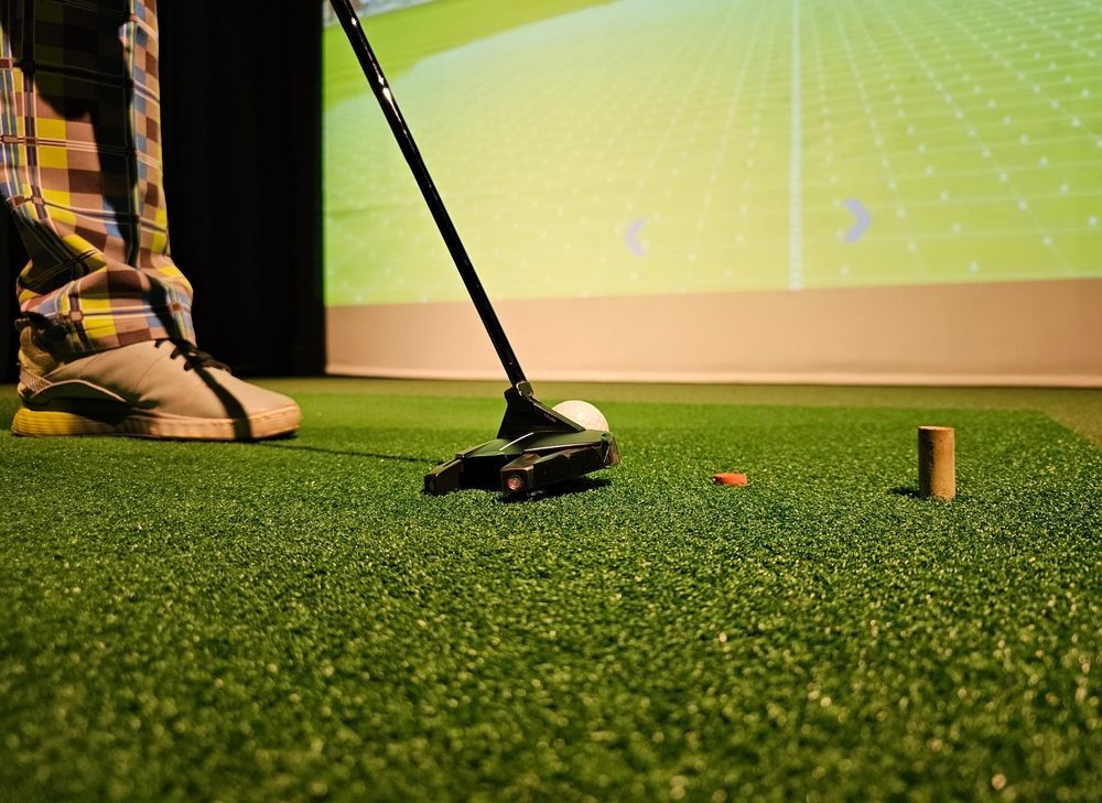 Golfer putting a golf ball on green turf, indoors; screen in background.