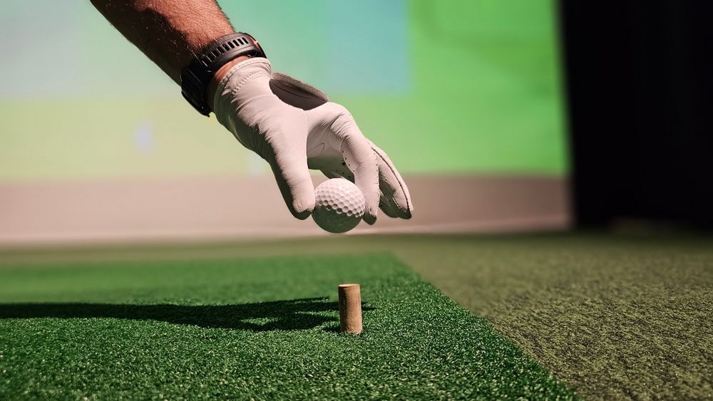 A gloved hand placing a golf ball on a tee on artificial turf.