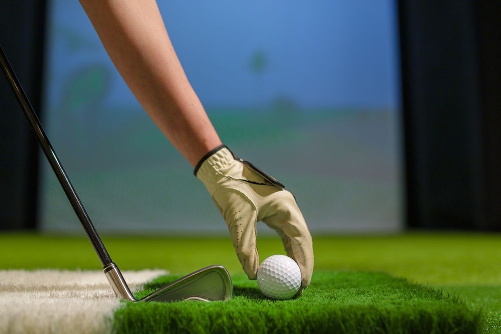Hand in golf glove placing a white golf ball on green artificial turf next to a golf club.
