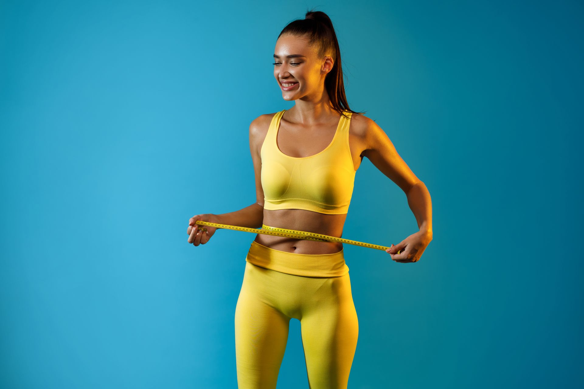 Woman in yellow workout clothes measuring her waist with a tape against a blue background