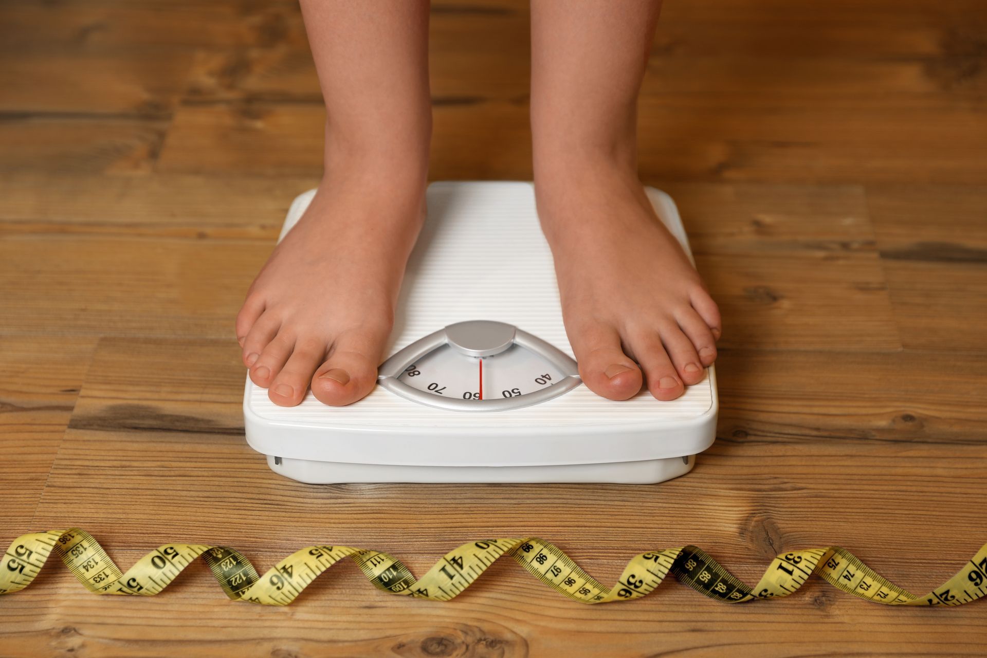 Feet on a bathroom scale beside a measuring tape on a wooden floor